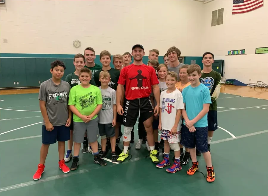 A coach in a red shirt standing with a group of young wrestlers on a gym mat