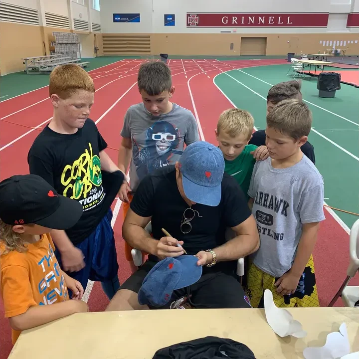 A group of young wrestlers gathered around an adult signing a cap at a camp