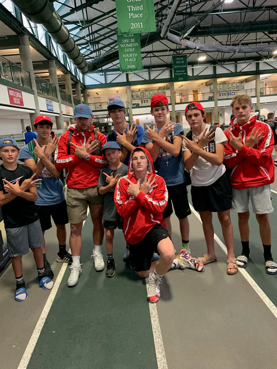 A coach and a group of young wrestlers, several in red jackets, posing on an indoor track